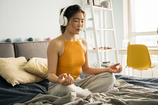Relaxed woman wearing headphones sitting at home in the bed in the lotus position.